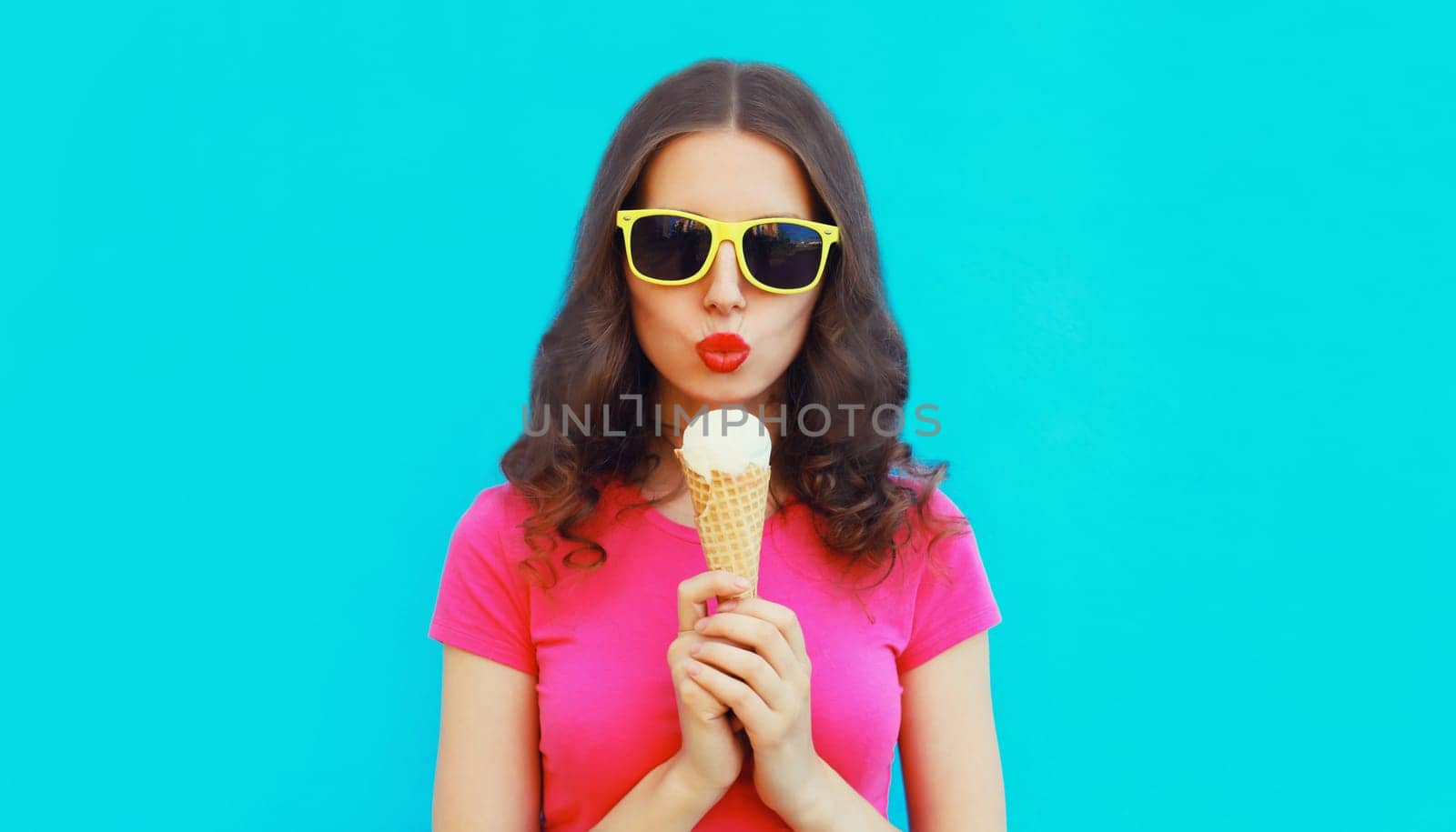 Summer portrait of happy young woman eating ice cream wearing sunglasses on blue background