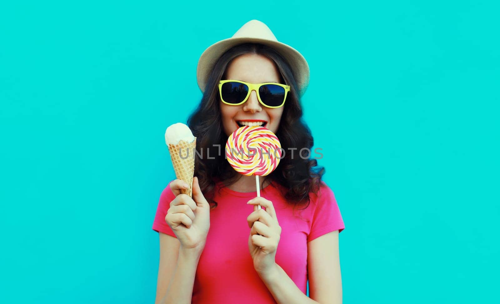 Summer portrait of happy young woman eating lollipop and ice cream wearing sunglasses and straw tourist hat on blue background