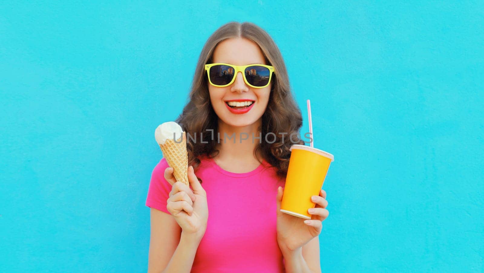 Summer portrait of happy smiling young woman eating ice cream and holding cup of fresh juice wearing sunglasses on blue background