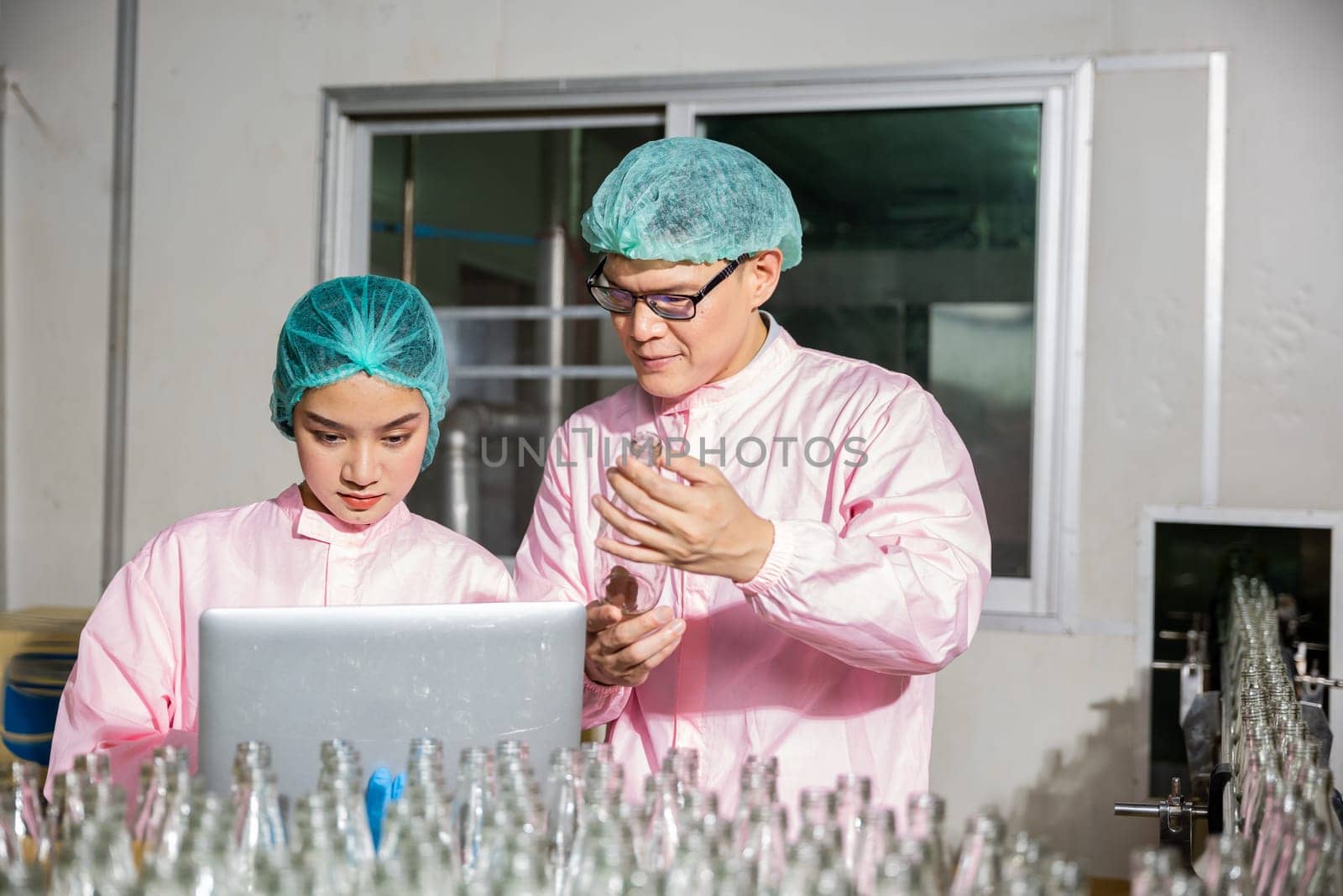 Two engineers conduct product bottle checks on a beverage factory's conveyor belt. Inspection and quality control handled by skilled professionals using a laptop for meticulous scrutiny.