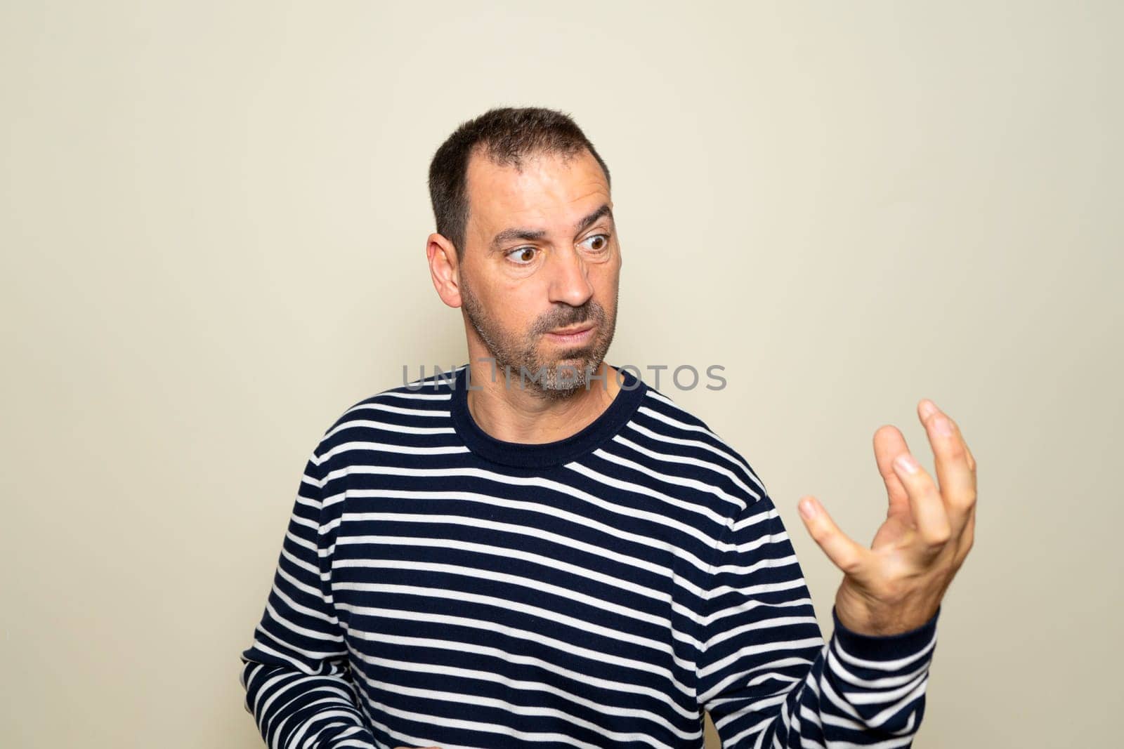 Bearded hispanic man in his 40s wearing a sweater looking extremely angry showing his claw hand while having an aggressive and defiant expression, isolated over beige background
