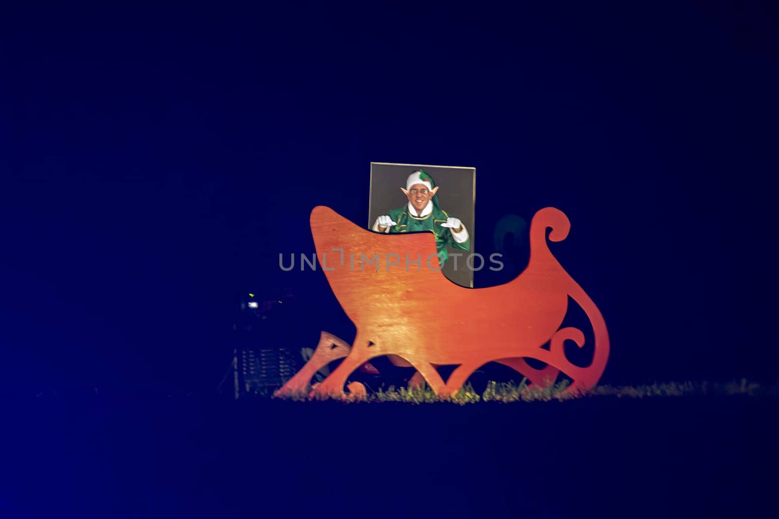 Nighttime View Of A Large Wooden Cut-Out Of Santa's Sleigh With A Person Posing As An Elf Through A Face-In-Hole Board, Creating A Festive Photo Opportunity.