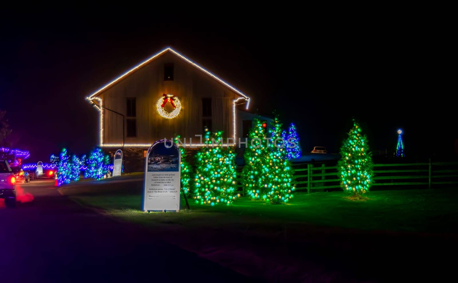 Festive Nighttime Scene Of A Building Outlined With White Christmas Lights And A Wreath, Surrounded By Trees Lit With Green Lights, Next To A Sign With Text And A Christmas Display.