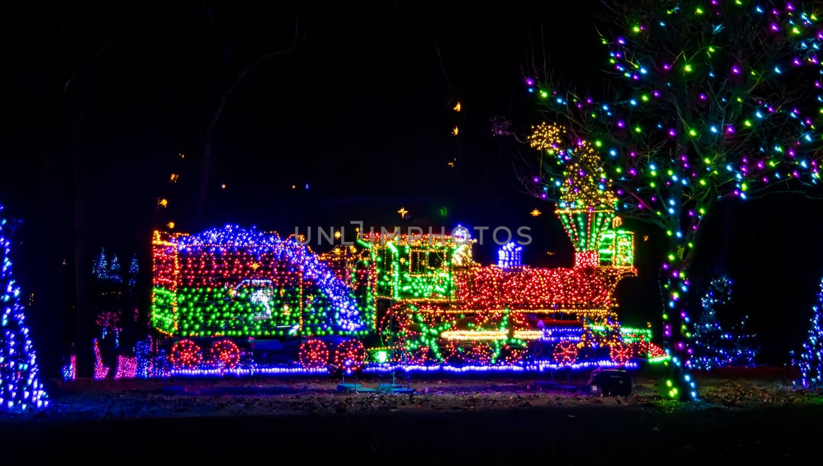 Night View Of An Elaborate Christmas Light Display Featuring A Multicolored Lit Train And Surrounding Trees, Creating A Vibrant And Festive Outdoor Scene.