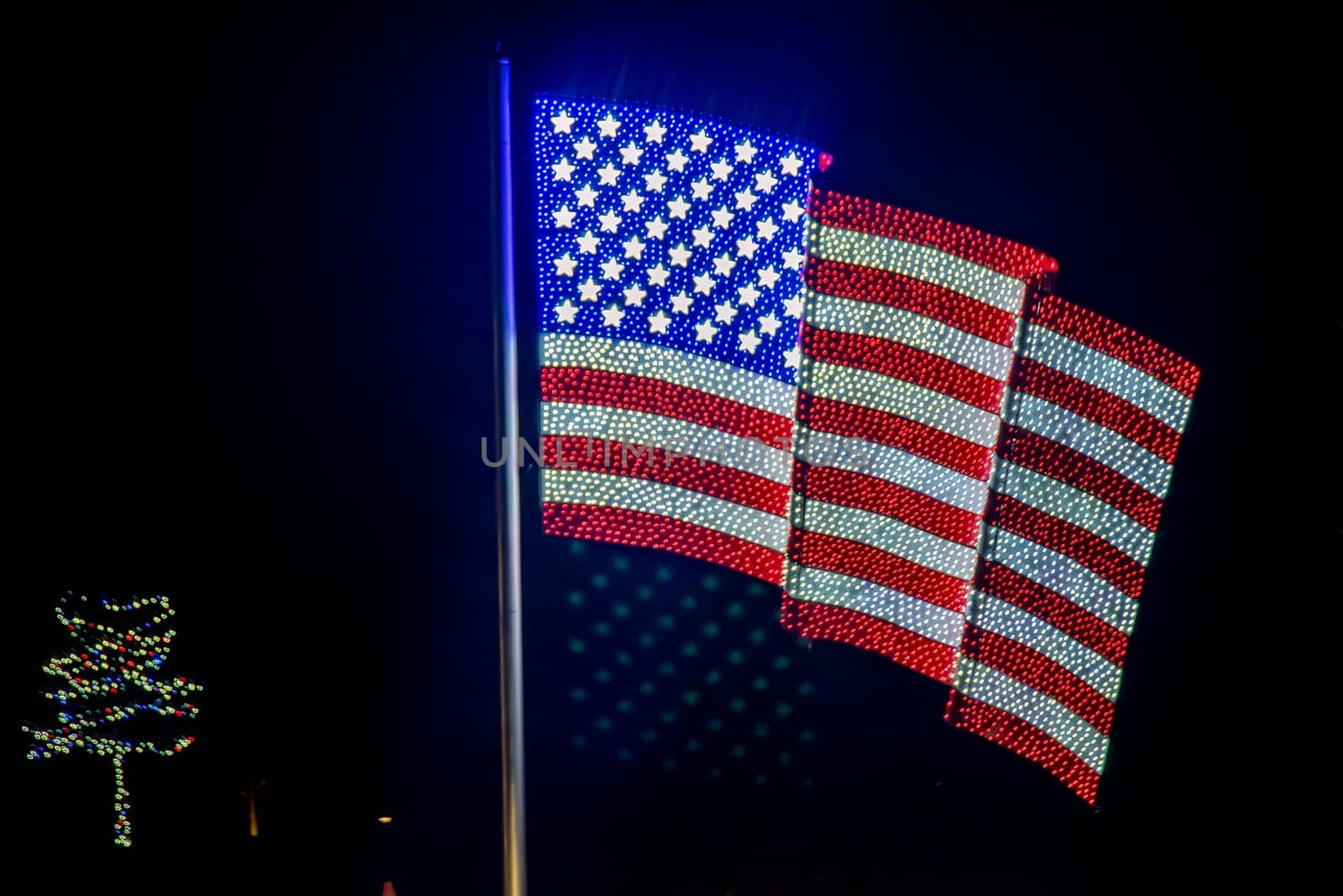 Nighttime Display Of Three American Flags Made Of Bright Lights Mounted On A Pole, With A Christmas Tree Light Display Visible To The Left Side.