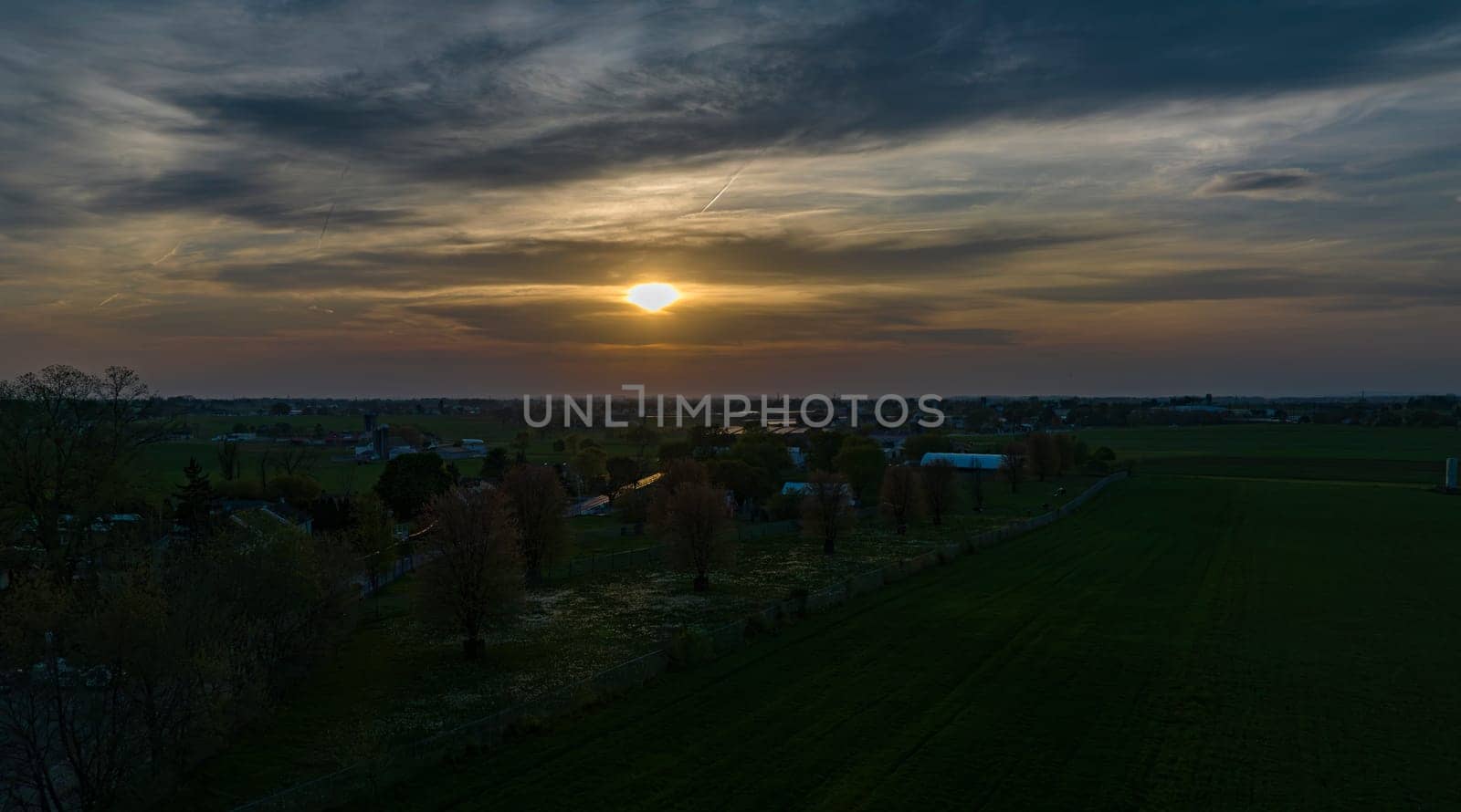 Dramatic Sunset Casting A Warm Glow Over A Rural Landscape With Silhouetted Trees And Buildings Under A Cloud-Streaked Sky.