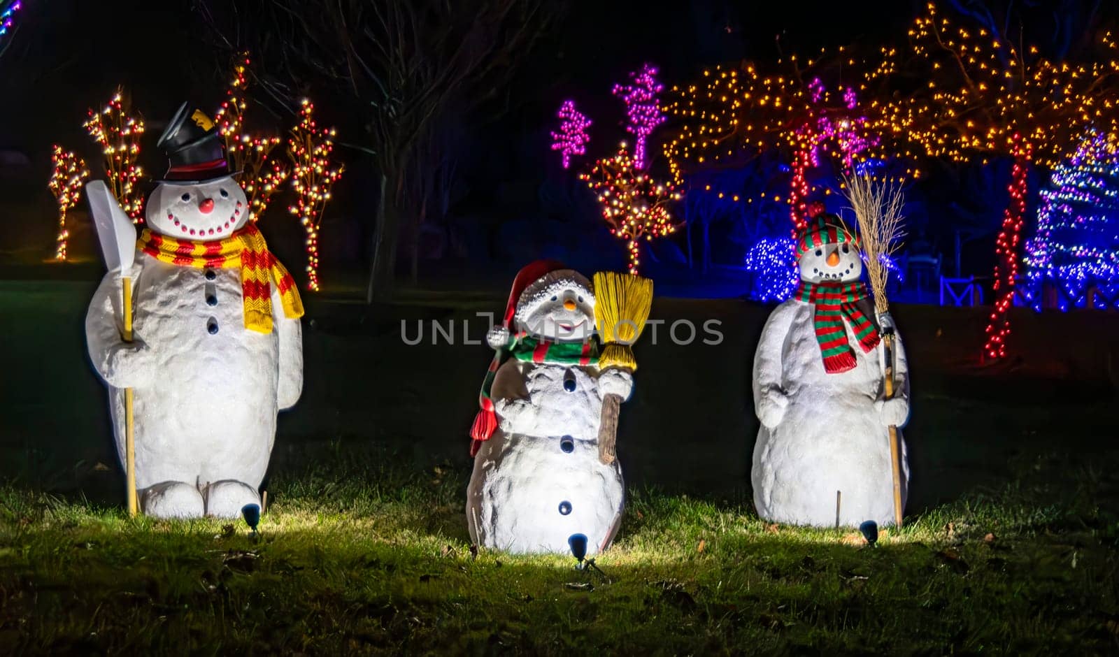 Three Illuminated Snowman Figures With Scarves And Hats Holding Brooms, Displayed At Night With Trees Decorated With Colorful Christmas Lights In The Background.