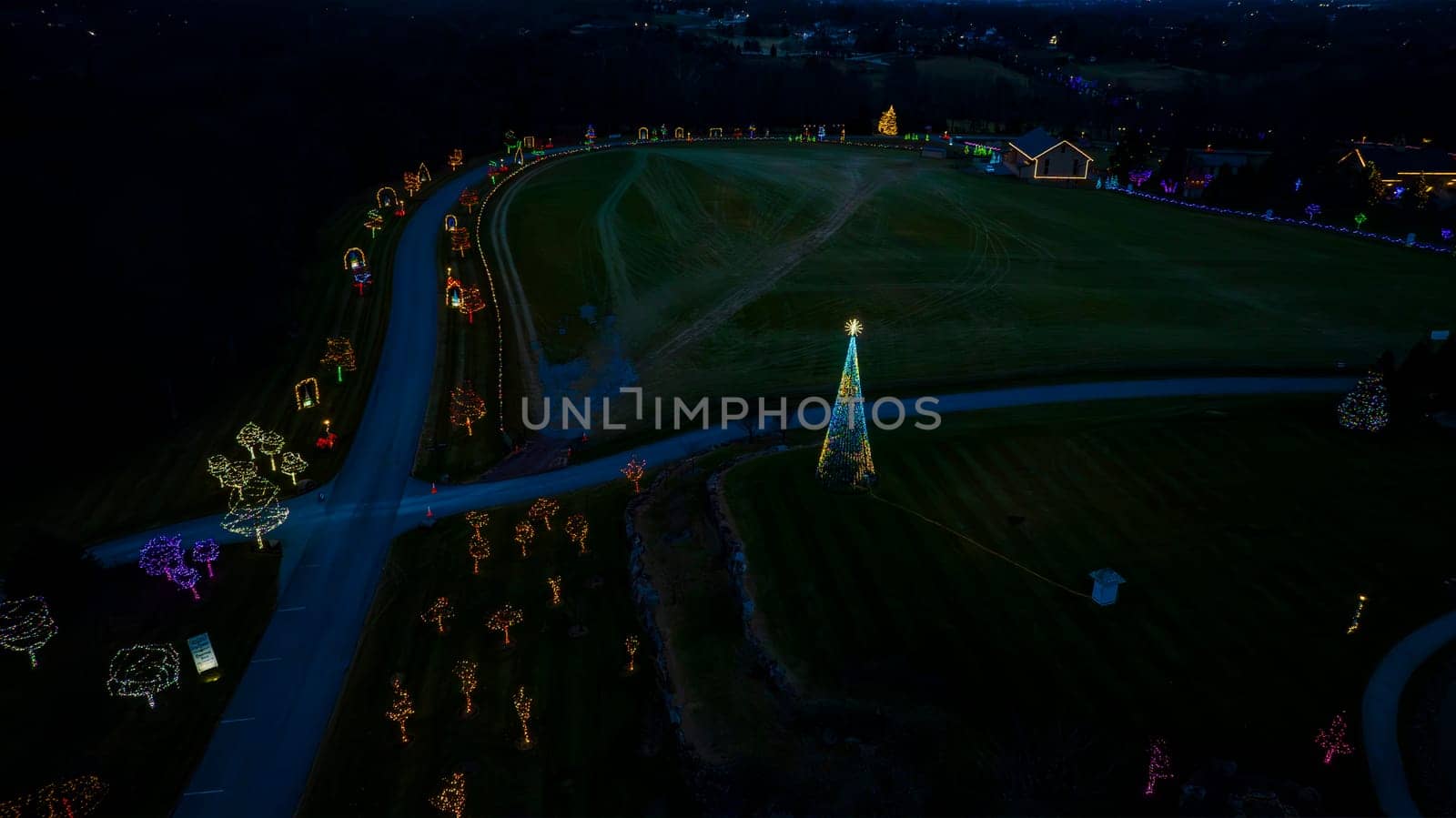 Aerial Nighttime View Of A Large Lit Christmas Tree And Multiple Smaller Trees Decorated With Colorful Lights Along A Winding Road.