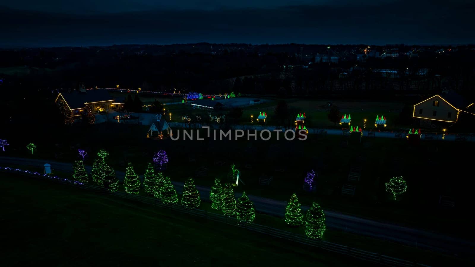 Elevated View Of A Festive Display With Two Houses Adorned In Christmas Lights And Surrounding Trees Decoratively Lit In A Rural Setting At Dusk.