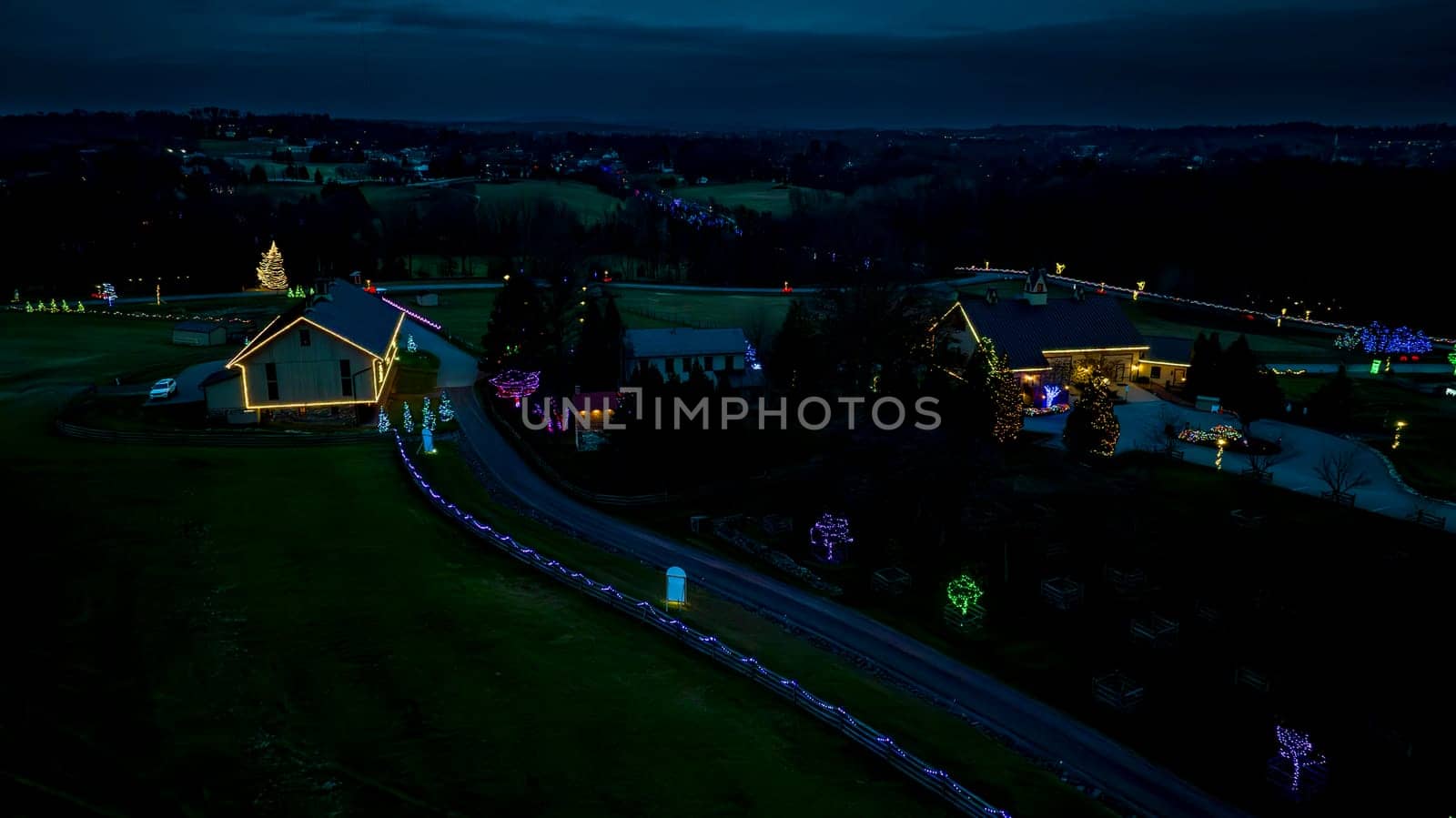 Nightfall Scene Over A Countryside Christmas Light Display With Illuminated Buildings And Trees Along A Curved Pathway.