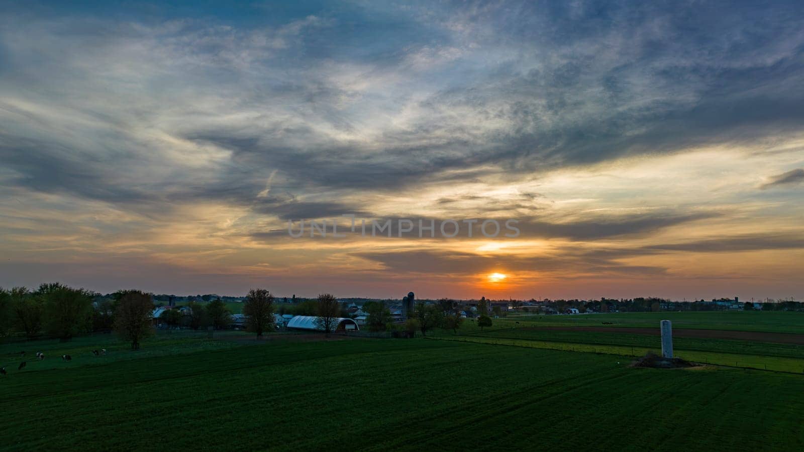 Aerial View of Dramatic Sunset Casting A Warm Glow Over A Rural Landscape With Silhouetted Trees And Buildings Under A Cloud-Streaked Sky.