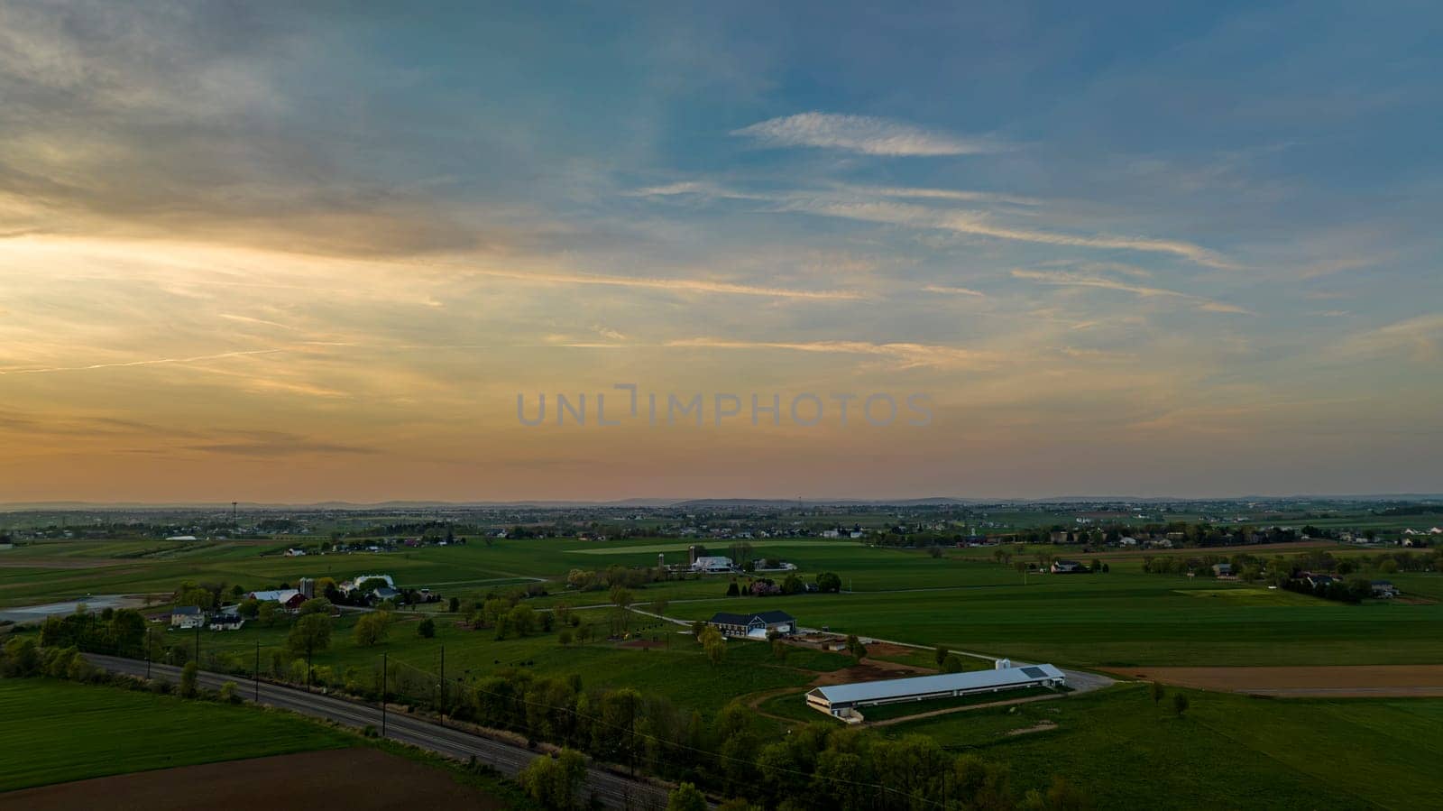 Aerial View of Dramatic Sunset Casting A Warm Glow Over A Rural Landscape With Silhouetted Trees And Buildings Under A Cloud-Streaked Sky.