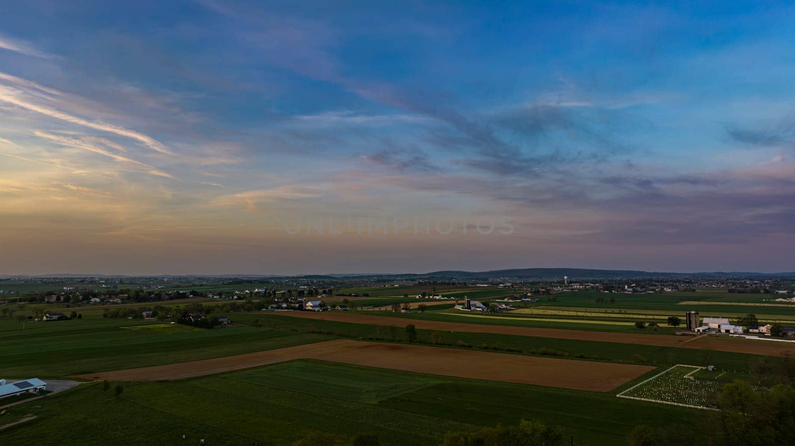 Expansive View Of A Rural Landscape At Twilight With Patchwork Fields, Sparse Buildings, And A Softly Colored Sky.