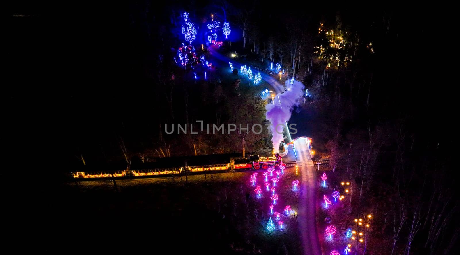 Overhead Night View Of A Colorful Light-Festooned Train Emitting Steam As It Travels Through A Dark, Tree-Lined Area.