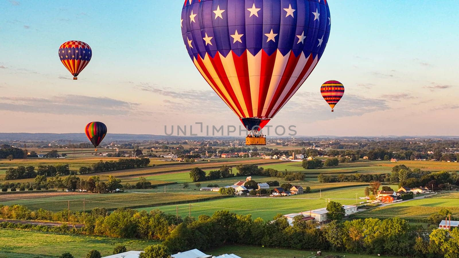 Patriotic-Themed Hot Air Balloon Dominates The Foreground With Others Dotting The Sky Above A Serene Rural Town As The Sun Sets.