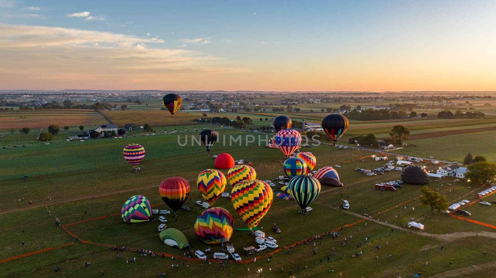An Aerial View of Cluster Of Colorful Hot Air Balloons Preparing For Flight At Dawn With Spectators And Vehicles Gathered On A Rural Field.