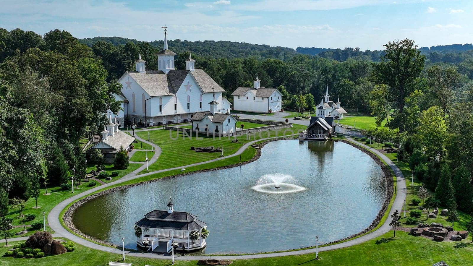 Aerial View Showcasing A Cluster Of Traditional White Orthodox Churches With Cross-Topped Domes, Arranged Around A Curved Pond With A Fountain, Amidst Green Trees And Neatly Arranged White Benches