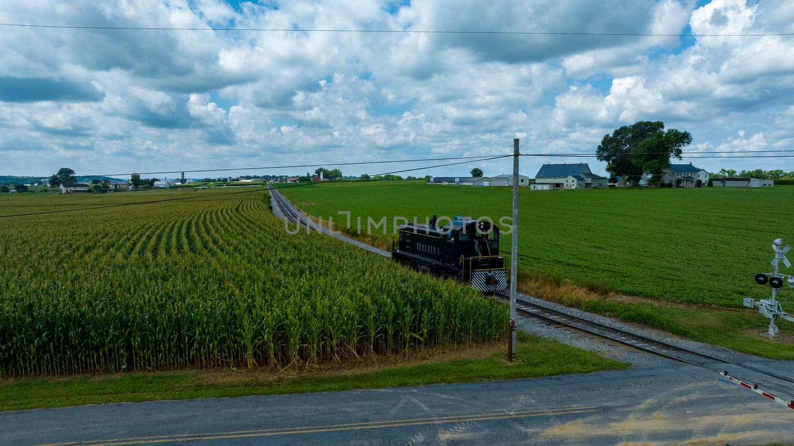 Dark Green Locomotive Moving On Tracks Beside A Cornfield With A Crossing Signal In The Foreground And Farm Buildings In The Distance Under A Dramatic Cloudy Sky.