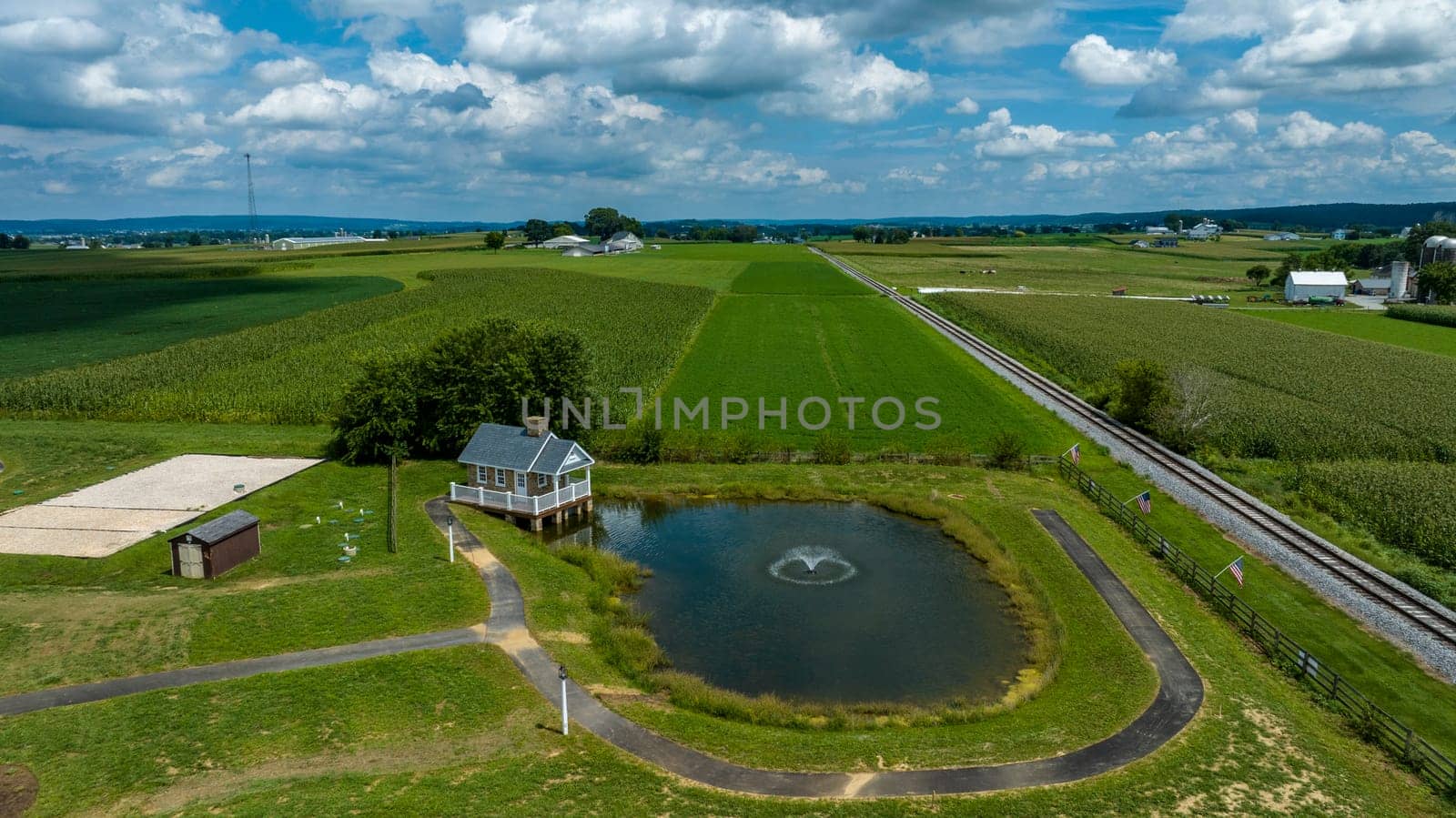 A Small Pond With A Water Fountain Is Situated Near A Curved Pathway And A Quaint House, With Parallel Railroad Tracks And Expansive Crop Fields Stretching Into The Distance Beneath A Cloudy Sky.