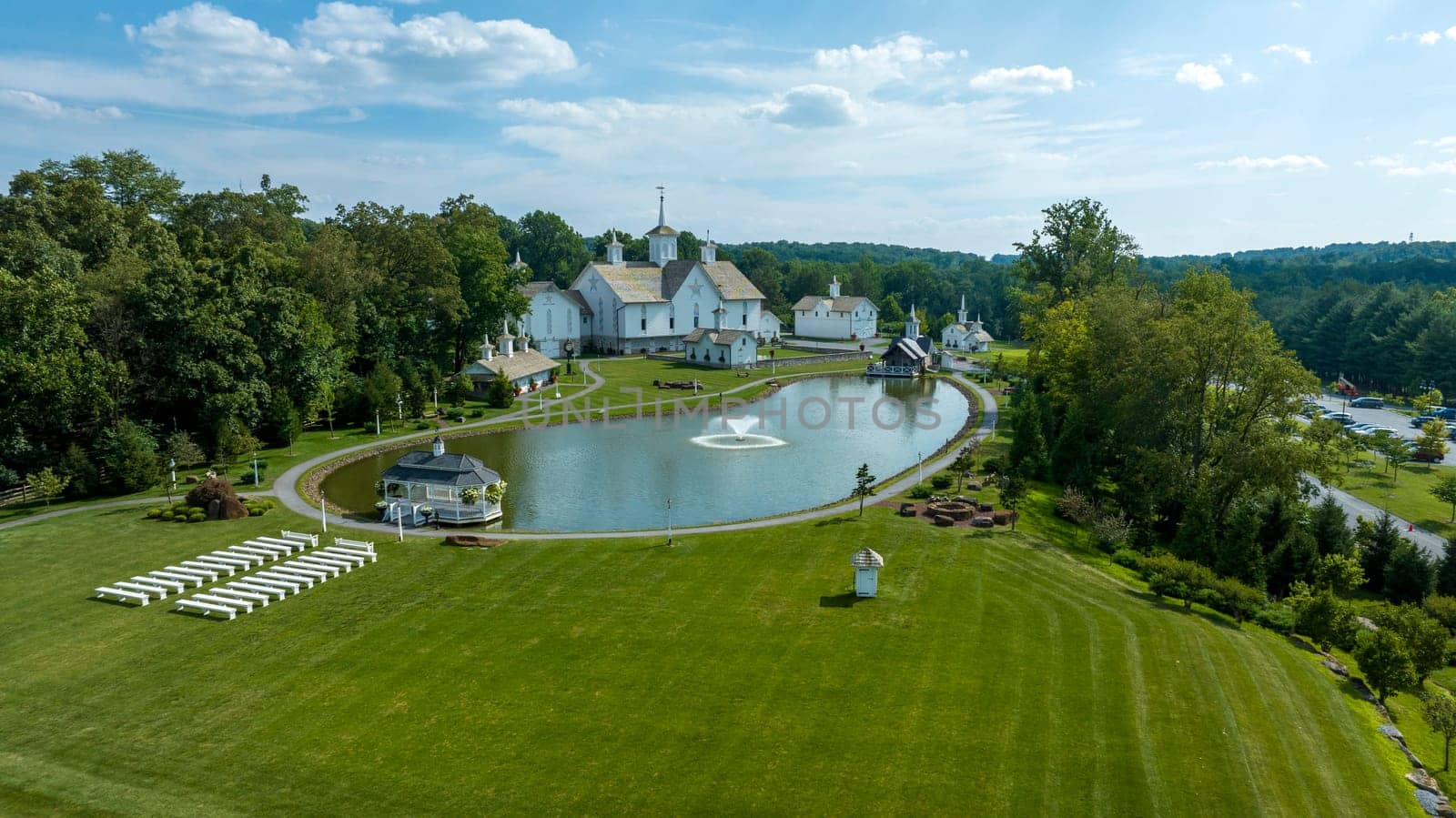 Aerial View Capturing A Series Of White Buildings With Orthodox Christian Architectural Features, Set Beside A Symmetrical Pond With Fountains, Flanked By Green Trees And Manicured Lawns.