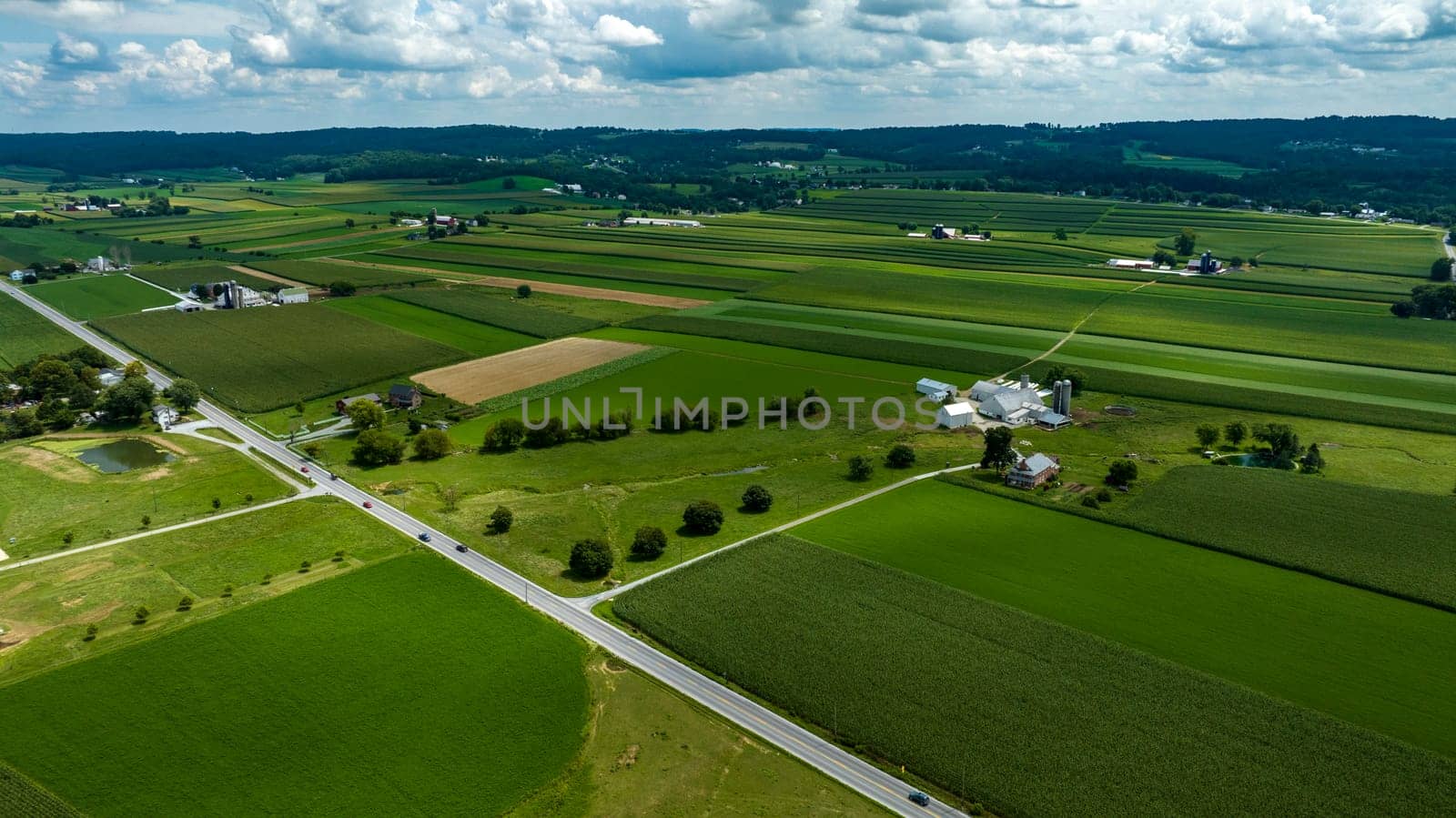 Aerial Perspective Of A Crossroads In A Verdant Rural Landscape With Patchwork Fields, Farm Structures, And A Pond Under Partly Cloudy Skies.