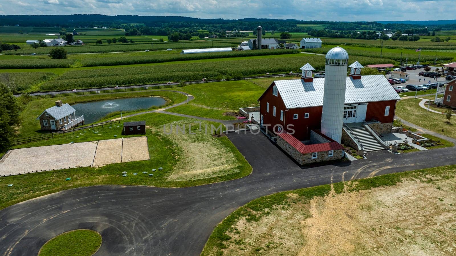 Overlooking A Farmstead Featuring A Majestic Red Barn With Silo, An Adjacent Pond With Geese, Winding Asphalt Paths, And Lush Green Crop Fields Under A Blue Sky With White Clouds.
