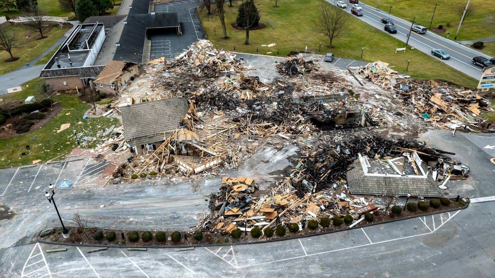 An Aerial View Of A Devastated Building Amidst Intact Structures With Cars On The Road And Green Fields In The Background Under A Cloudy Sky.