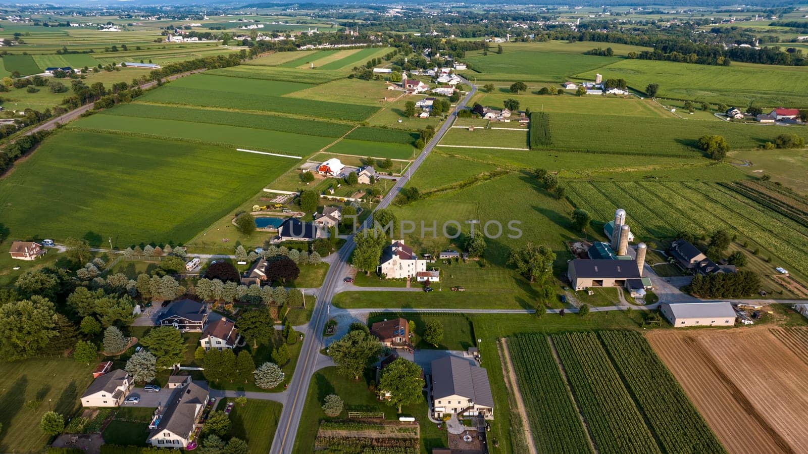 High-Angle View Of A Rural Community With Farms, Crops In Neat Rows, Roads Crisscrossing The Landscape, And Buildings Clustered Along The Thoroughfare.