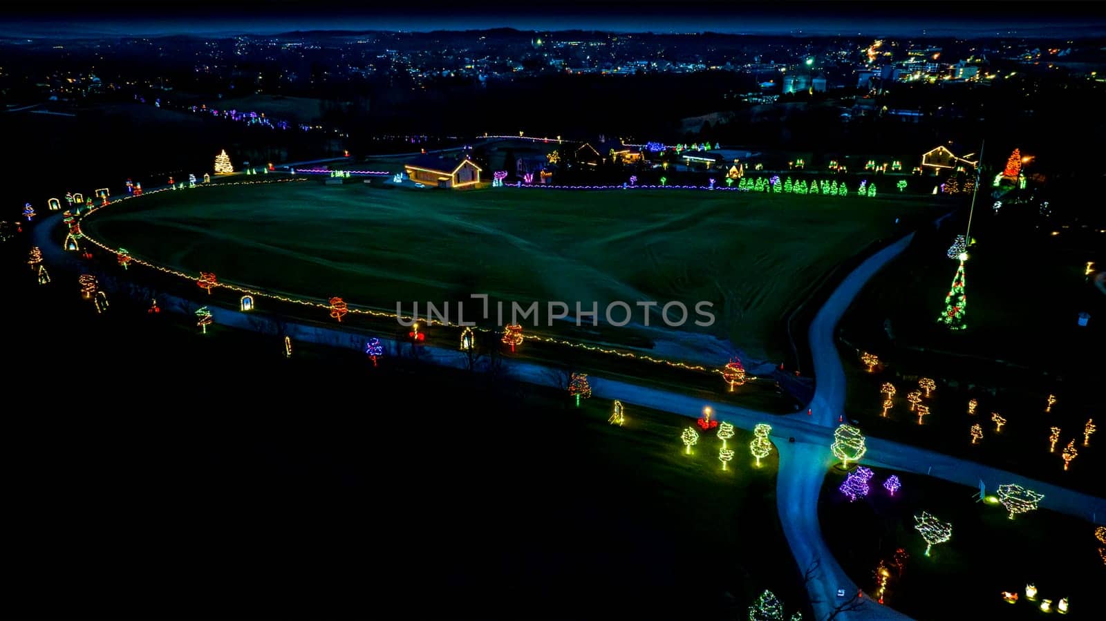 Aerial Night View Of A Winding Road Adorned With Luminous Christmas Trees Leading To A Centrally Lit Large Tree And Buildings.