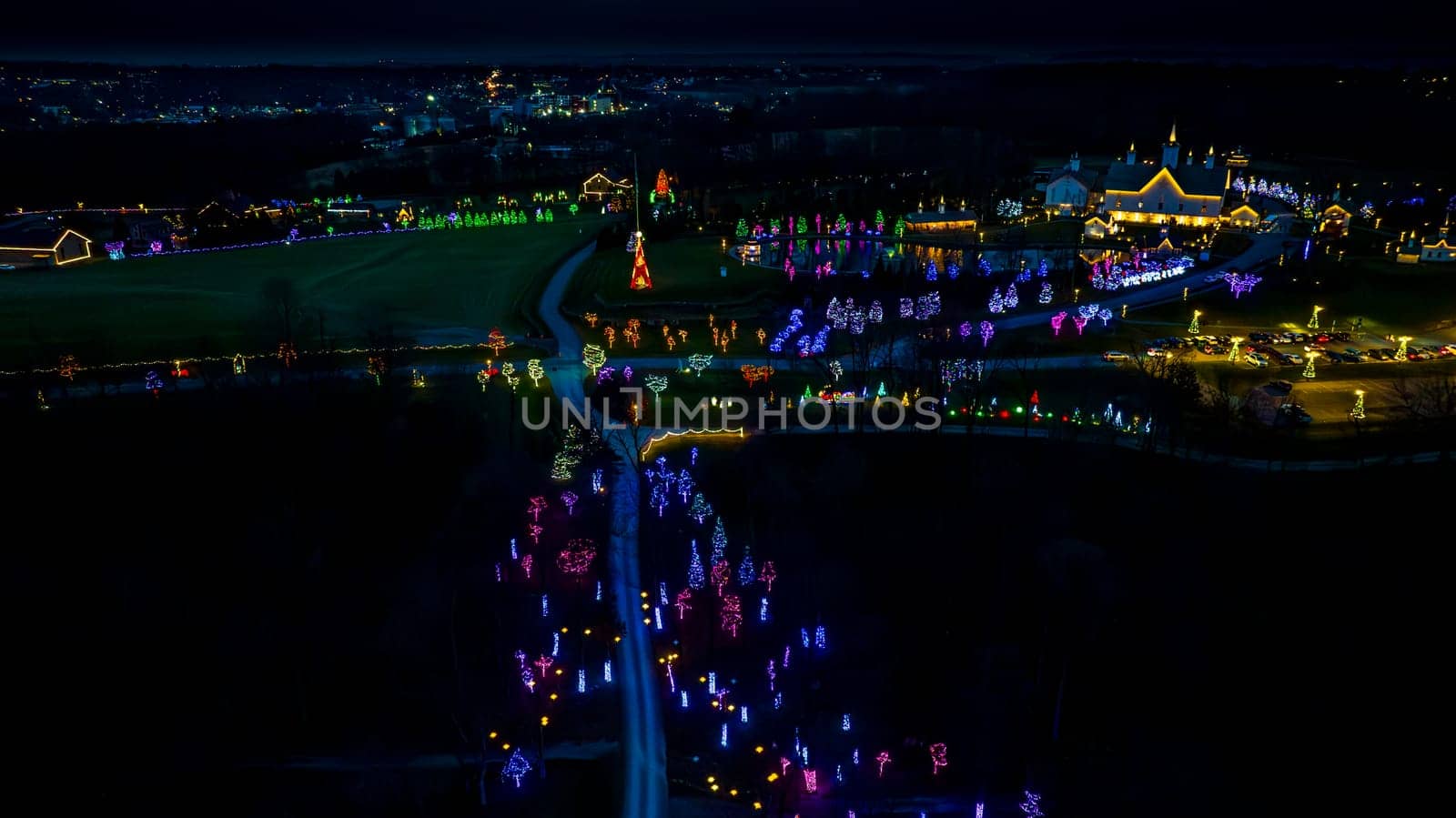 High-Angle Night View Of A Lively Holiday Lighting Display With A Road Leading To A Central Pond And Clustered Lit Structures.