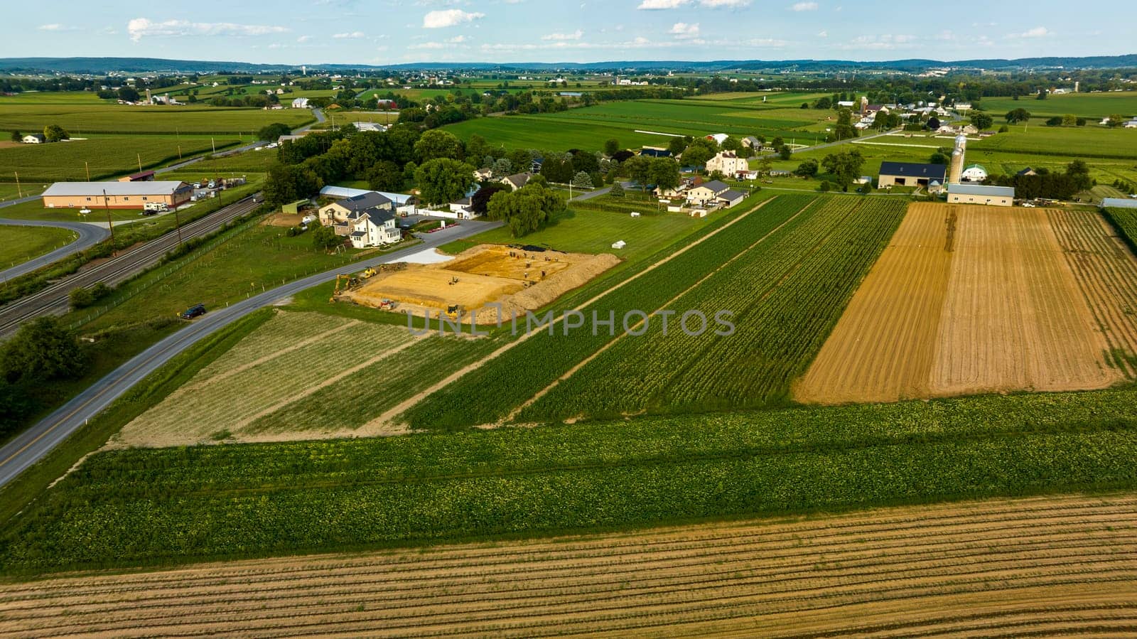 Aerial View Featuring Roads Intersecting Near Construction Site Amidst Farmland With Fields In Various Stages Of Cultivation And A Small Community In The Background.