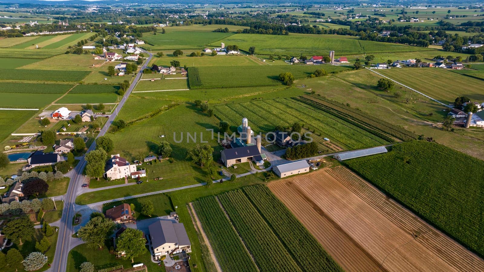 High-Angle View Of A Rural Community With Farms, Crops In Neat Rows, Roads Crisscrossing The Landscape, And Buildings Clustered Along The Thoroughfare.