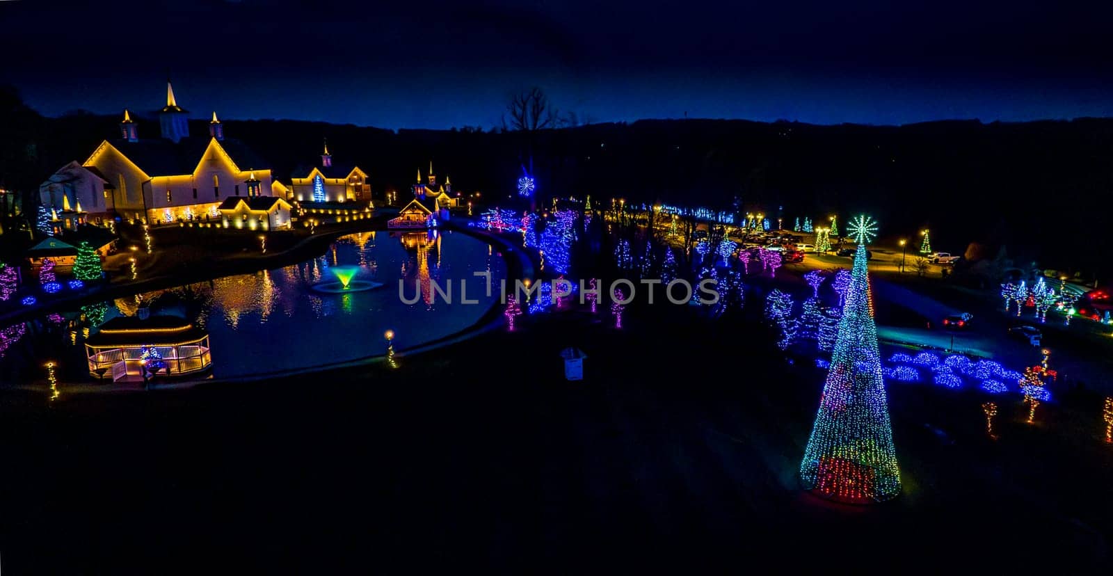Aerial View Of A Grand Holiday Lights Display Featuring A Reflective Pond, A Giant Christmas Tree, And Buildings With Illuminated Roofs At Night.