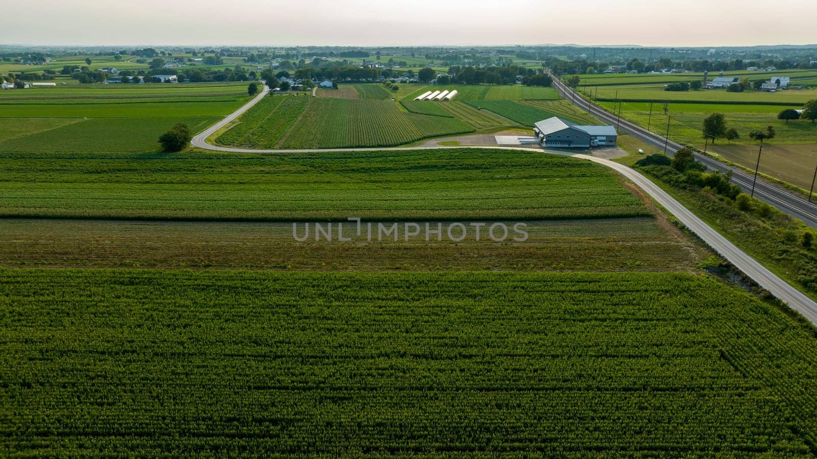 High Angle View Showcasing A Stretch Of Road And Railroad Tracks Cutting Through A Lush Tapestry Of Agricultural Fields With Farm Buildings In The Distance.