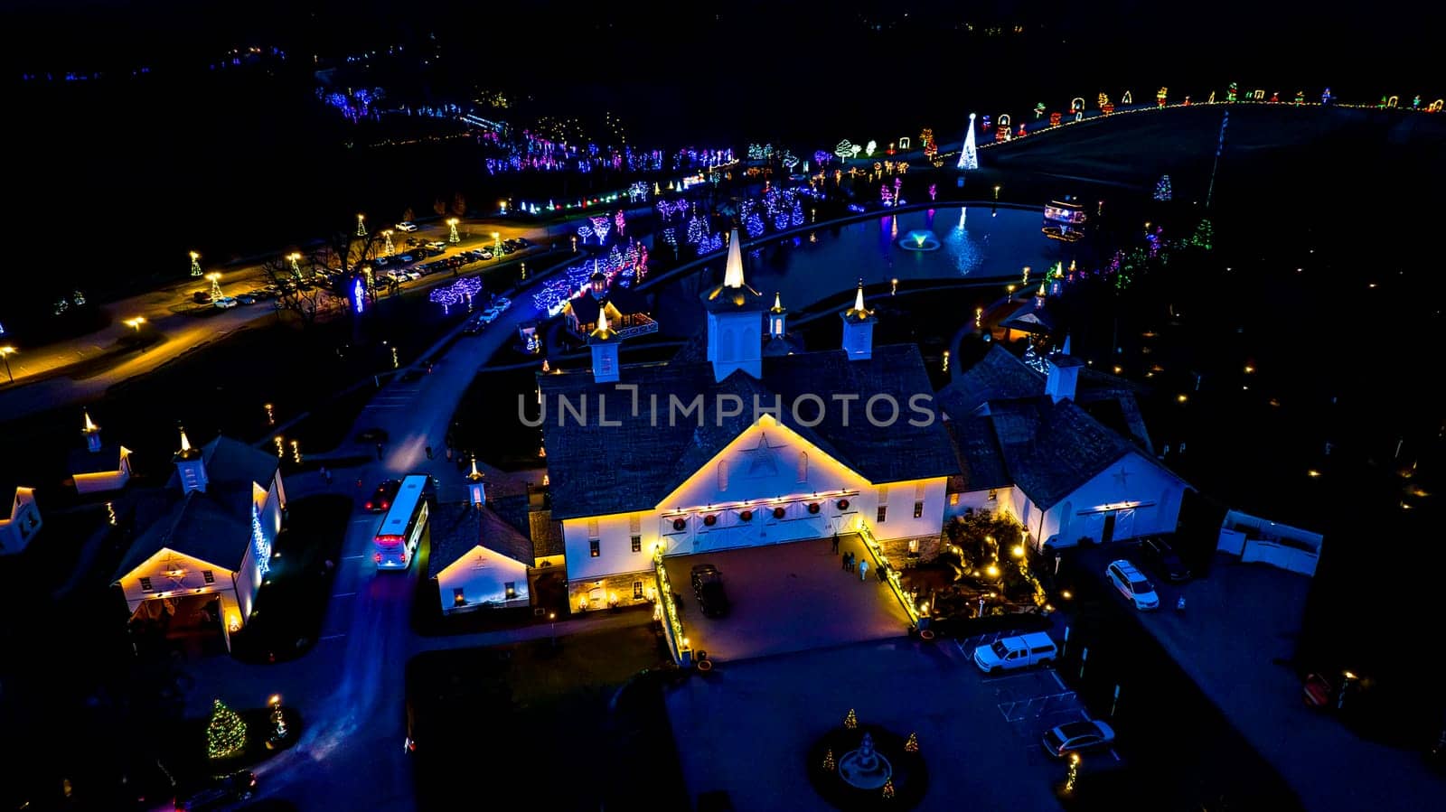 Overhead Night View Of A Complex With Buildings Outlined In Lights, A Central Pond, And A Surrounding Drive Lined With Lit Trees.