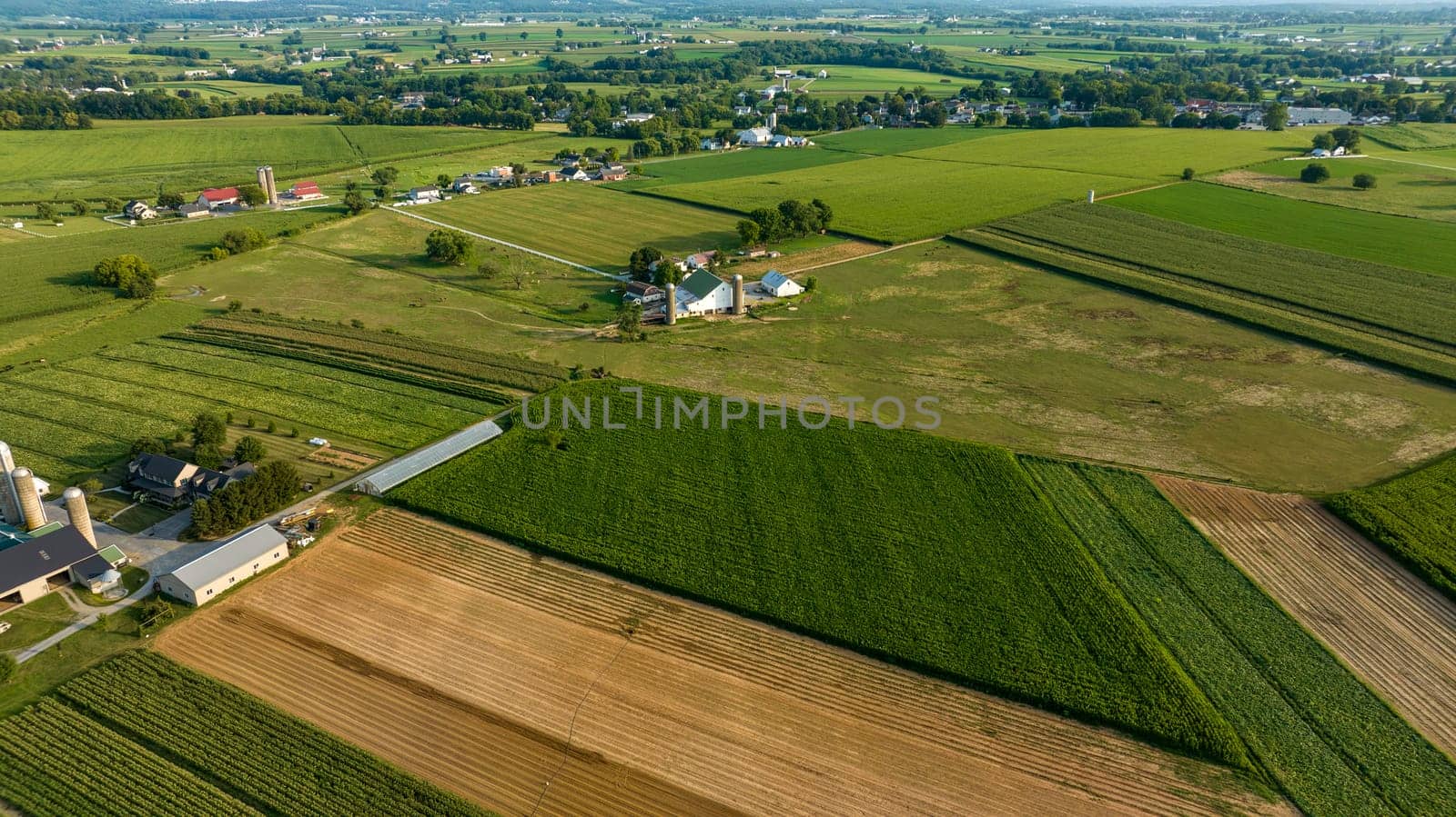 Diagonal Aerial View Of Varied Farmland With Different Crops, Farm Buildings, Silos, And A Network Of Roads In Early Evening Light.