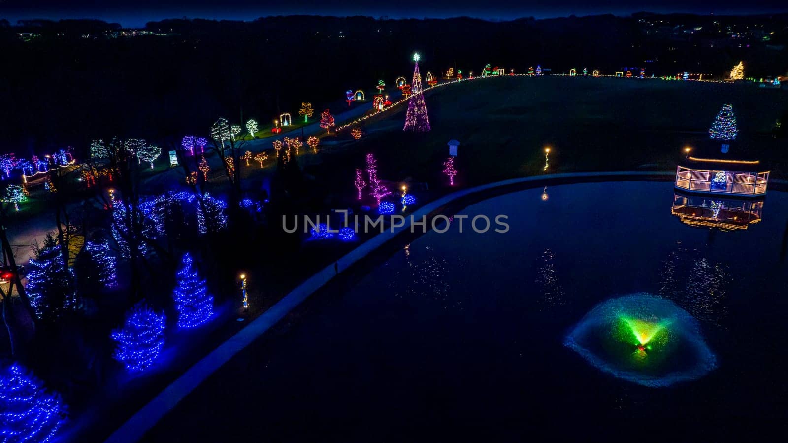 Overhead Nighttime View Of A Serene Pond With A Lit Fountain, Surrounded By A Curved Pathway With Festive Trees In Multicolored Lights.
