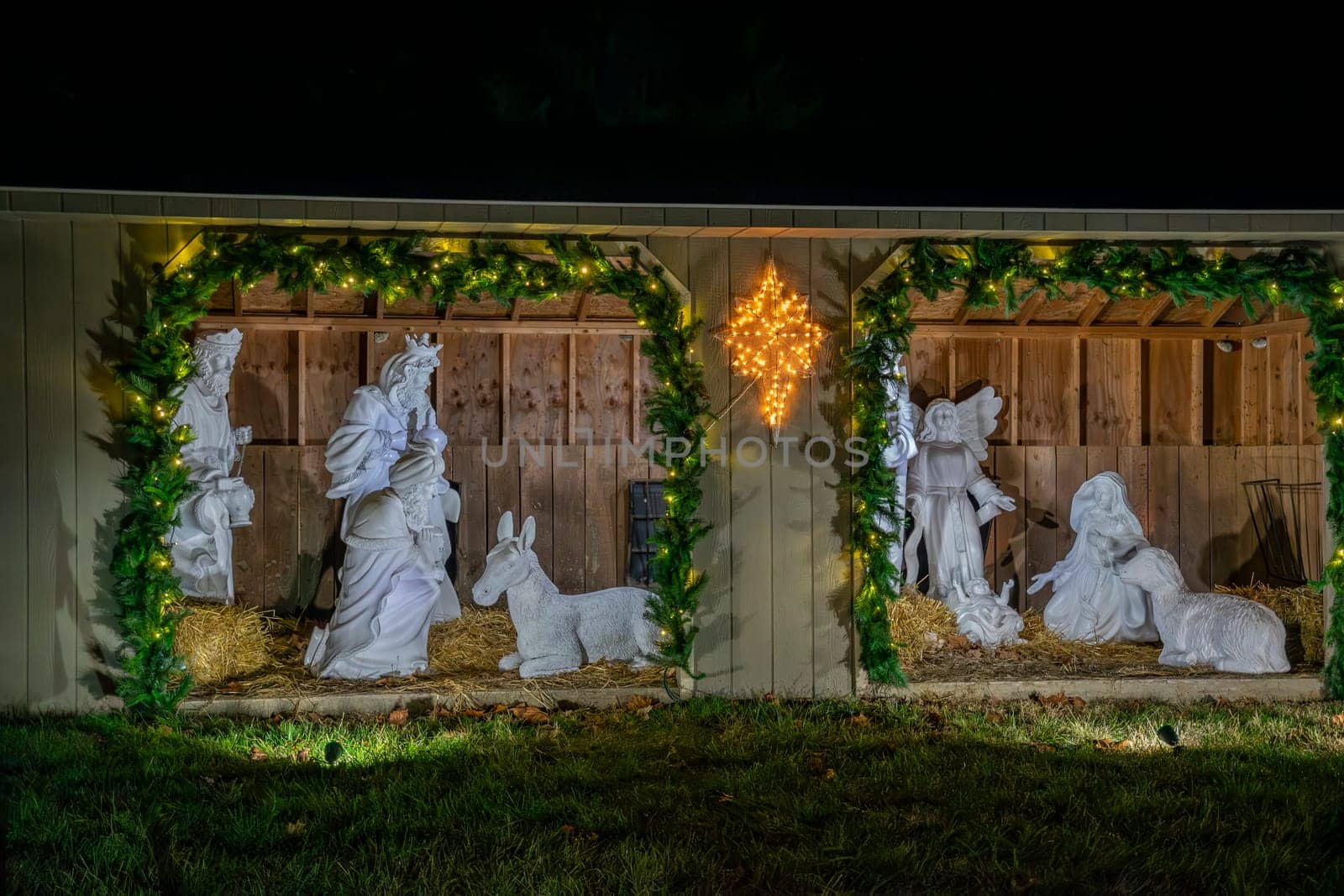 Nighttime Image Of A White Nativity Scene With Figures Of Mary, Joseph, And Wise Men Flanked By Animals, Illuminated By A Star And Garland Lights.