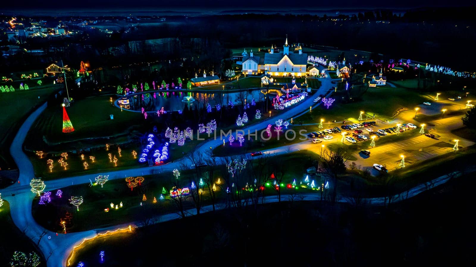 Expansive Aerial View Of A Festive Grounds Illuminated With Holiday Lights Featuring A Path To A Pond And Multiple Buildings At Night.