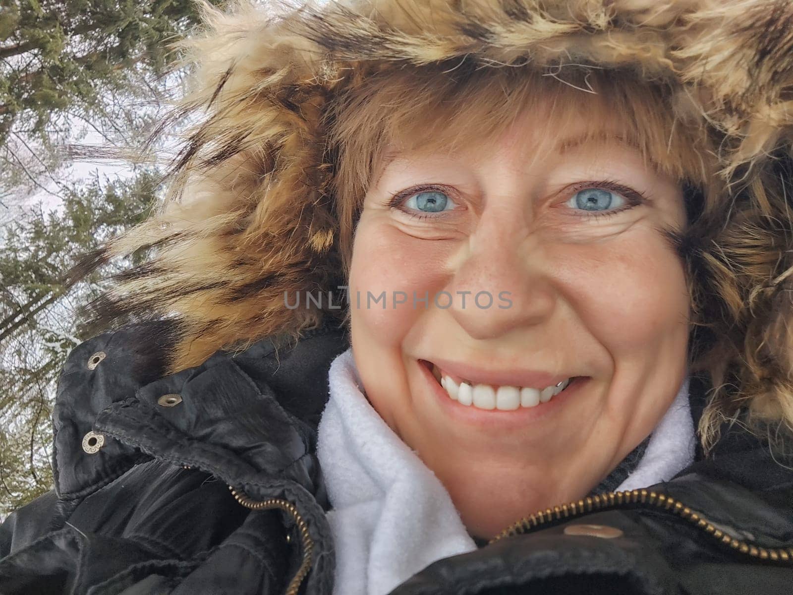 Happy, cheerful middle-aged woman in a warm jacket and a hood with fur taking selfie in winter outdoors. Female Traveler in Alaska, Lapland, Arctic, Siberia