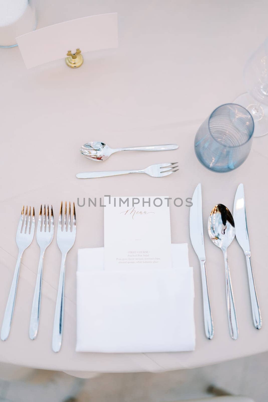 Personalized invitation lies in a white napkin on the table next to the cutlery, opposite a blank card on a stand by Nadtochiy