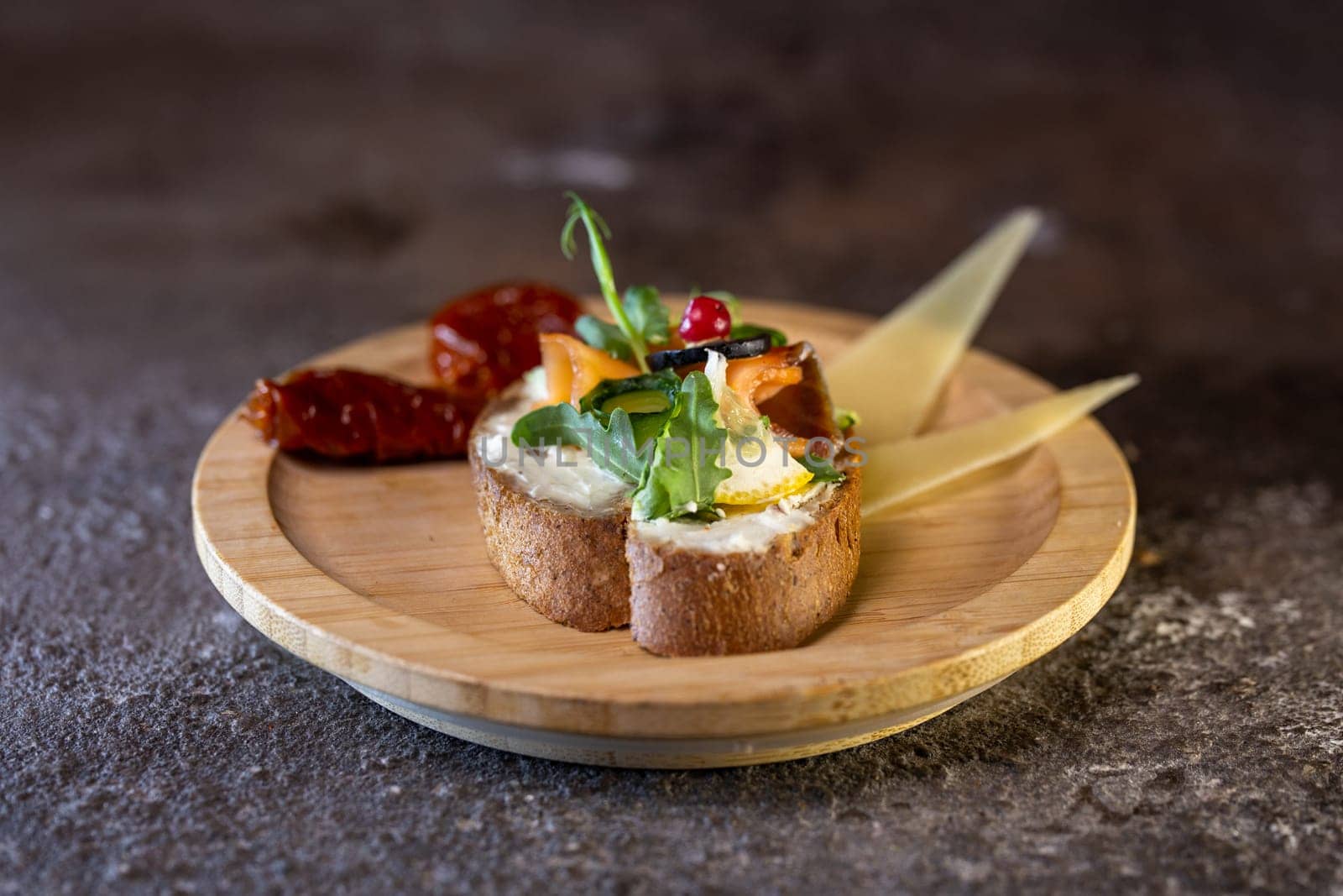 Wooden plate with bread, cheese, veggies, and greens on a dark background. Bread topped with cream cheese, veggies, and greens. Cheese in slices. Plate on wooden table.