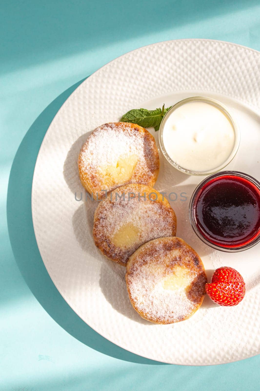 Four cheese pancakes with sour cream and strawberry jam on a white plate on a blue background. A delicious and healthy breakfast or dessert. Perfect for a food blog or recipe website.