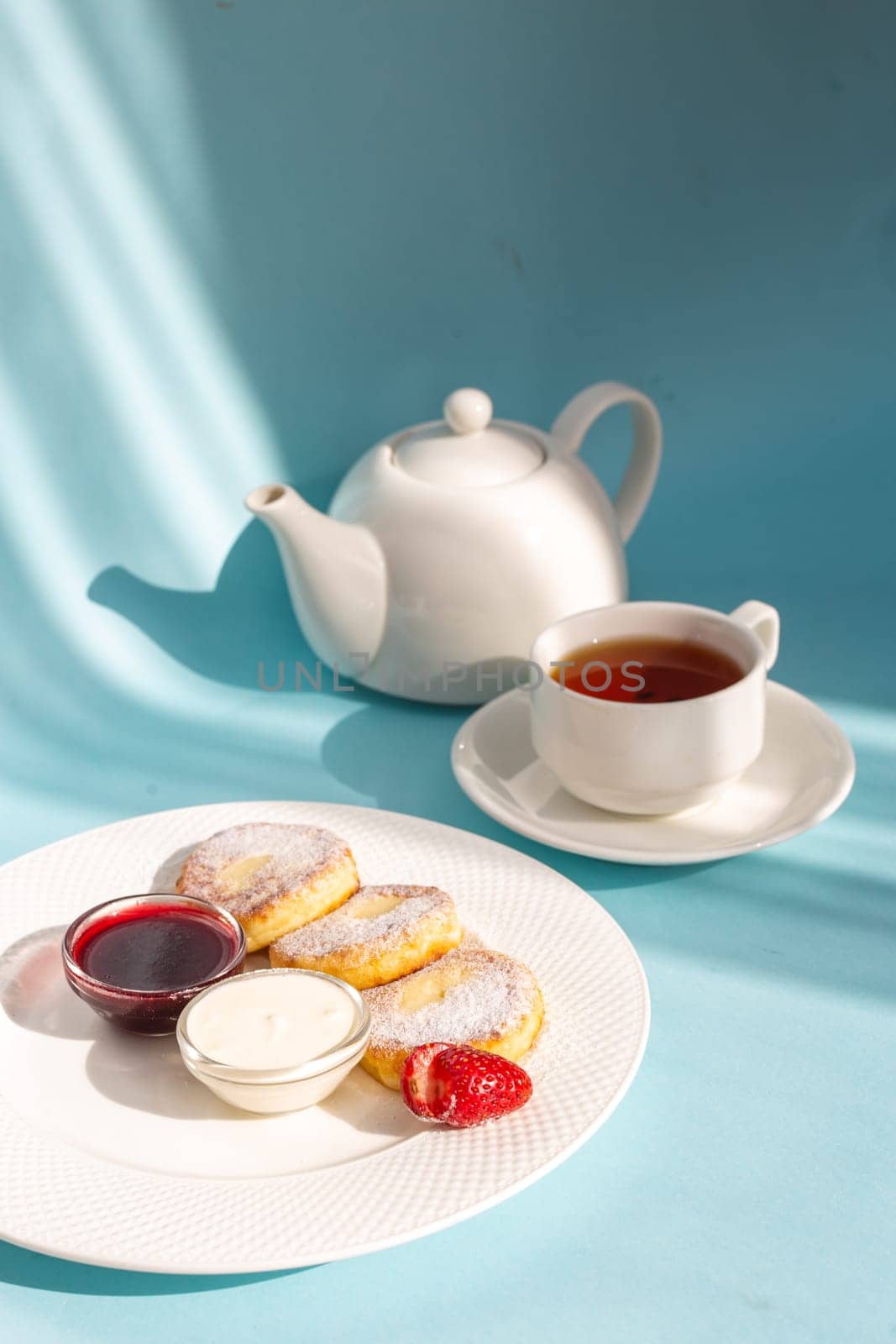 White plate with cottage cheese pancakes, sour cream, jam, and a cup of tea on a blue table. by Pukhovskiy