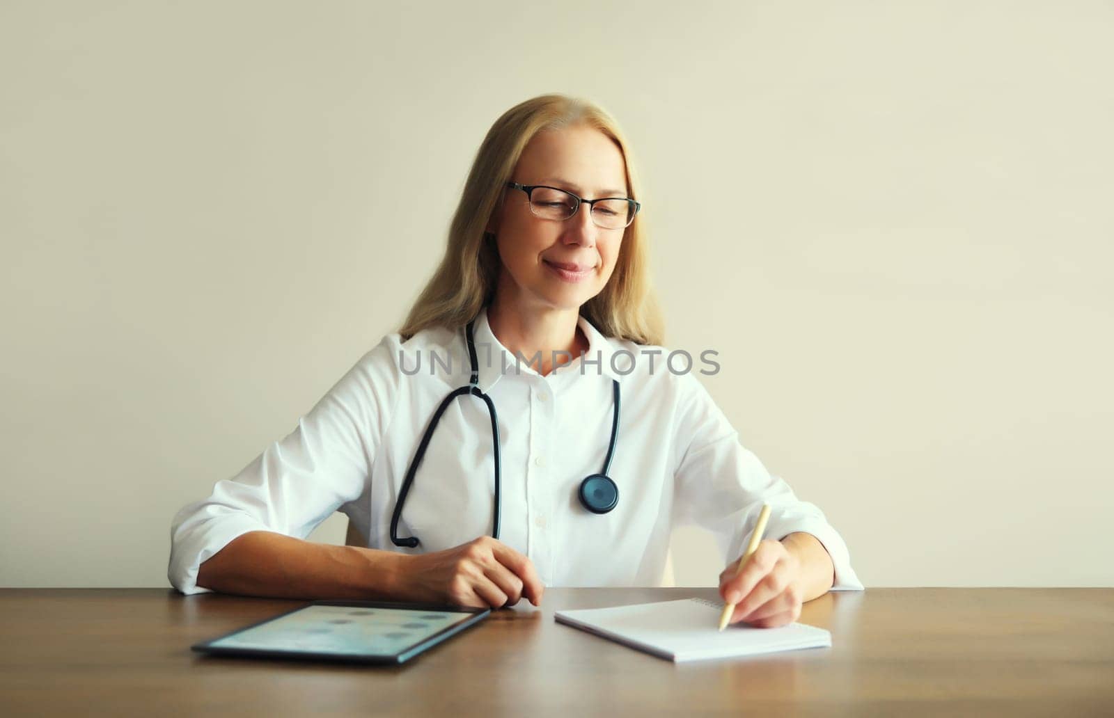 Caucasian middle-aged woman doctor with stethoscope, paper documents and digital tablet computer sitting at desk in clinic office