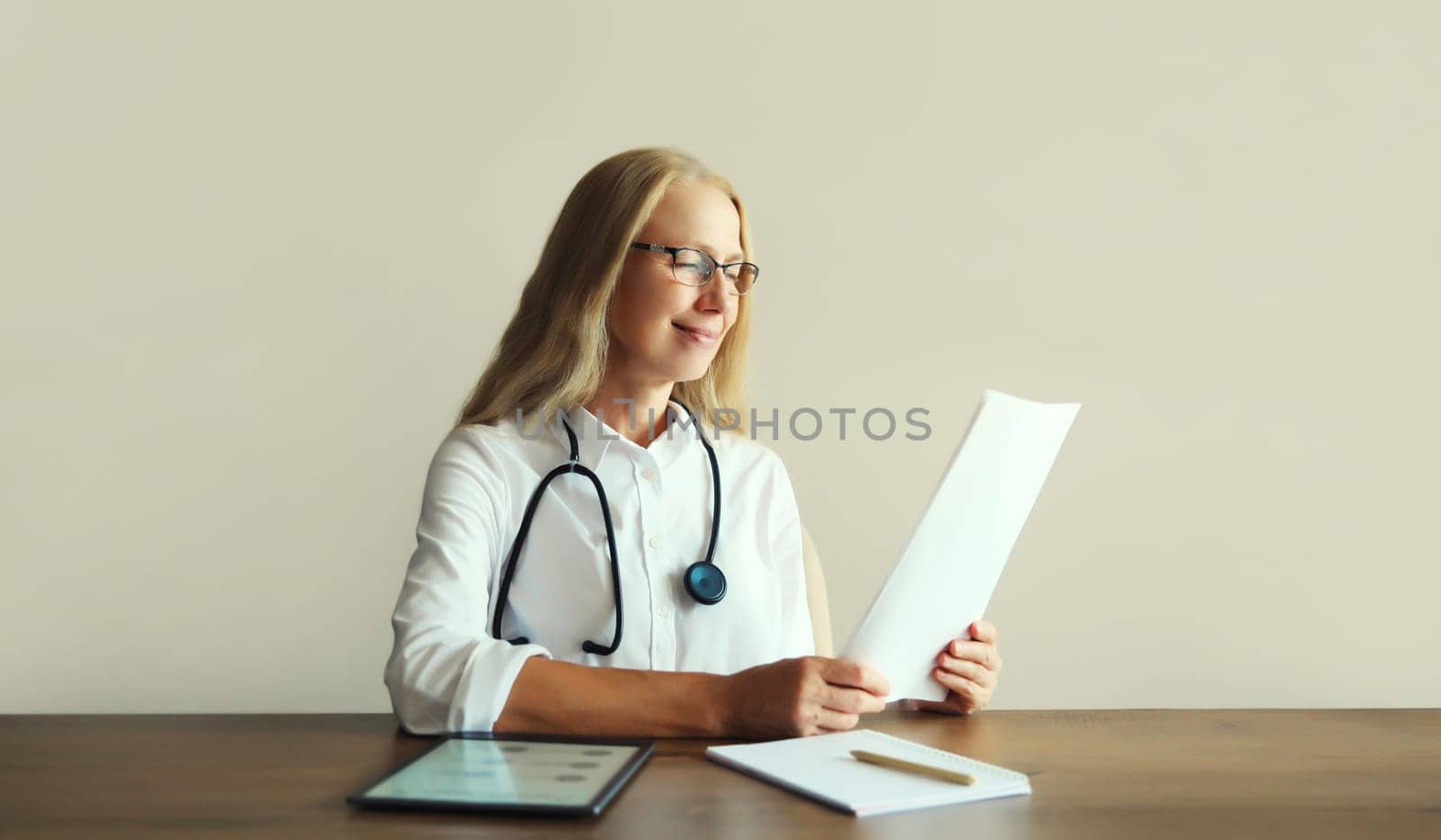 Caucasian middle-aged woman doctor with stethoscope, paper documents and digital tablet computer sitting at desk in clinic office