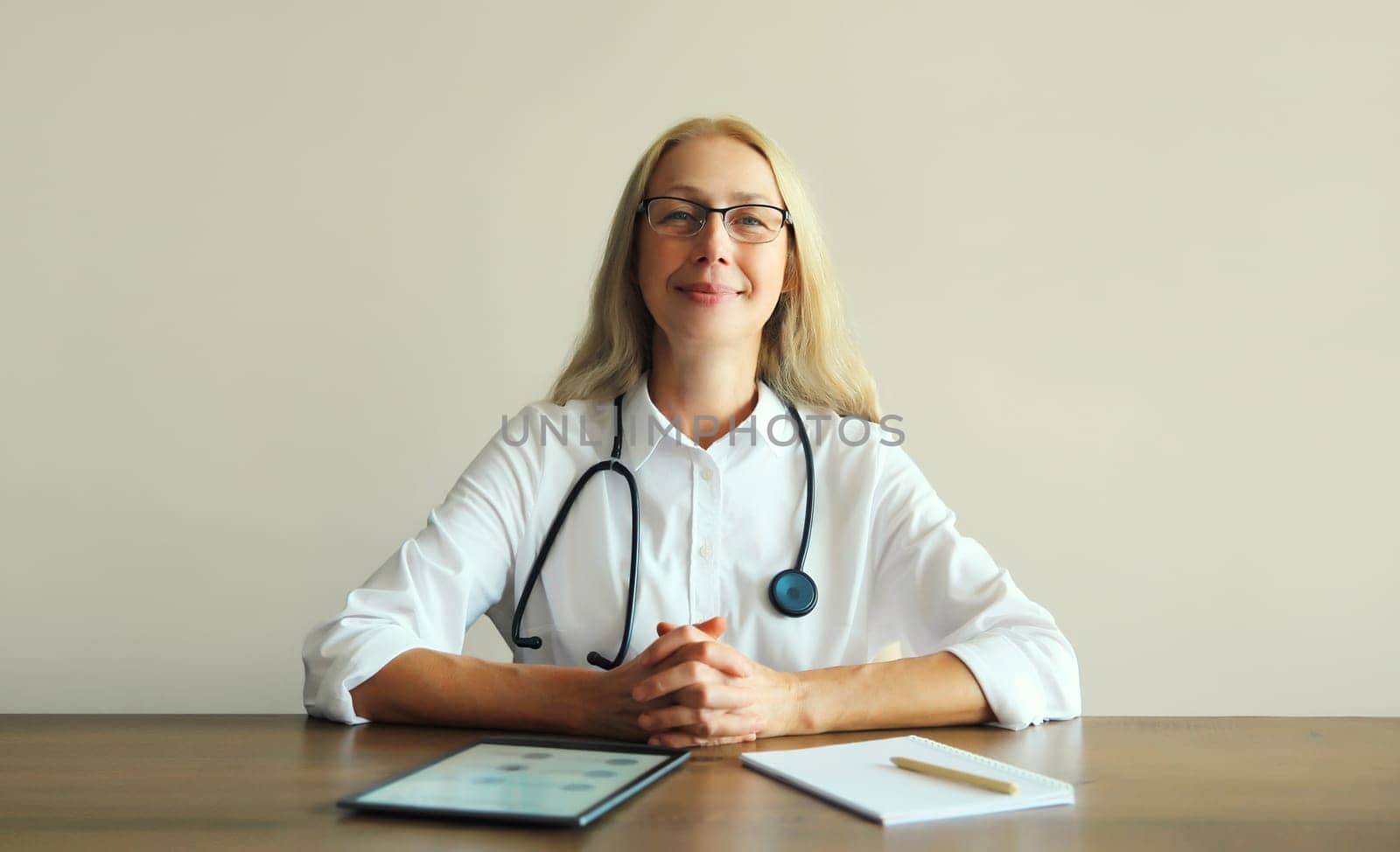 Caucasian middle-aged woman doctor with stethoscope, paper documents and digital tablet computer sitting at desk in clinic office