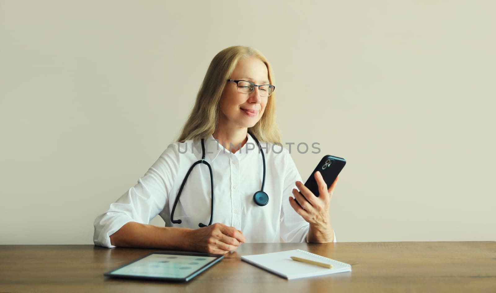 Caucasian middle-aged woman doctor using mobile phone with paper documents and digital tablet computer sitting at desk in clinic office