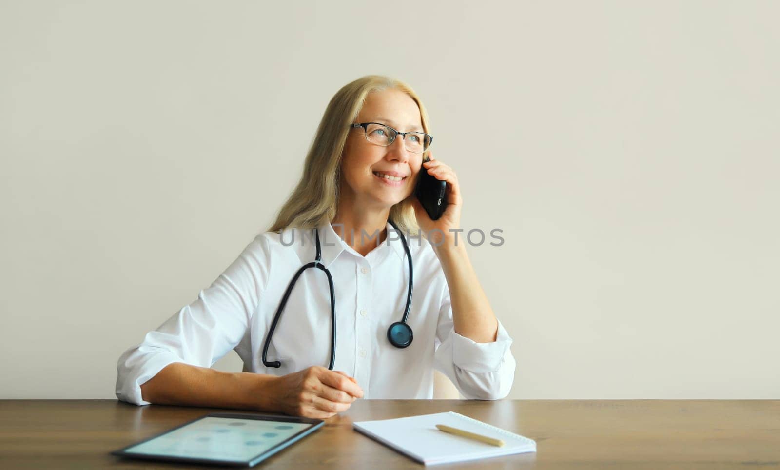 Caucasian middle-aged woman doctor calling on mobile phone with paper documents and digital tablet computer while sitting at desk in clinic office