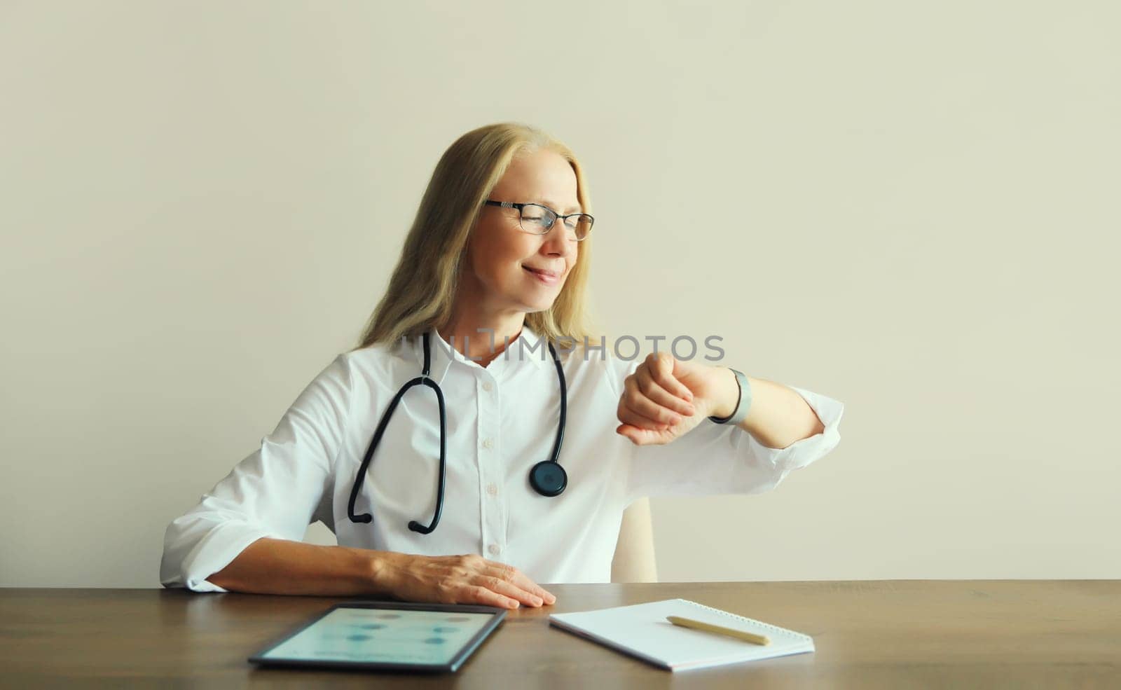 Caucasian middle-aged woman doctor with stethoscope, paper documents and digital tablet computer sitting at desk in clinic office